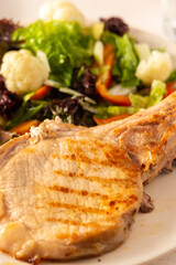 Fried pork entrecote on the bone with stewed pumpkin pieces and salad in a plate close up, green salad with cauliflower and napa cabbage, bell pepper and spices