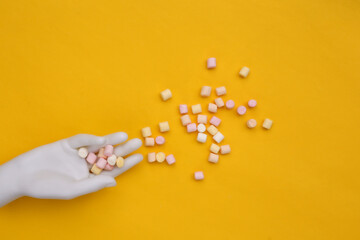 A lot of mini marshmallows in a white mannequin hand on a yellow background. Top view