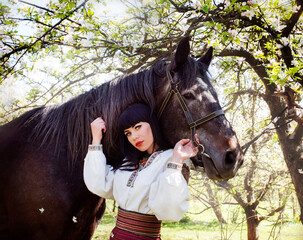 a girl touches her hands to a flowering tree in the garden. against the background the lodge grazes. Ukrainian national costume. brunette with bangs with plump lips hold the horse by the harness.