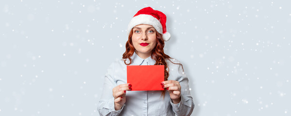 Portrait of cheerful girl in santa hat holding a red wishlist letter mockup with snow on light blue background. New year celebration banner, copy space.