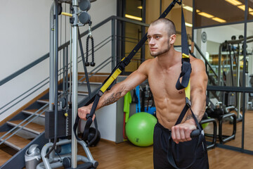 Muscular naked torso man training with fitness trx system in the gym. Bodybuilder exercising his muscles with suspension straps