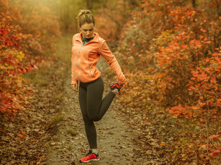 Young attractive sport woman in sportswear doing stretching leg in autumn forest with reddened leaves of trees. Warm up before running. Healthy lifestyle concept