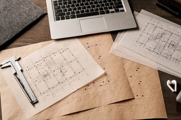 Laptop, handtool, pen and cordless earphones surrounded by unfolded blueprints