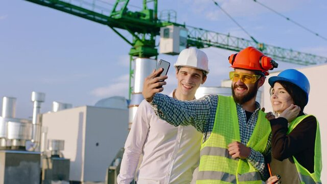 Closeup to the camera group of charismatic diverse specialists on the top of construction site wearing safety helmets taking some pictures using the smartphone