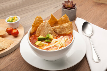 Mexican soup with tortilla chips, avocado and cheese in a white bowl on a rustic wooden table
