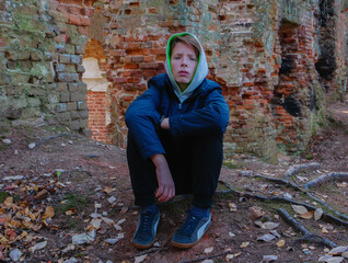 A little boy 12-15 years old sits near the old ruins of a red brick house. Teenager sits on the ground near a brick wall.
