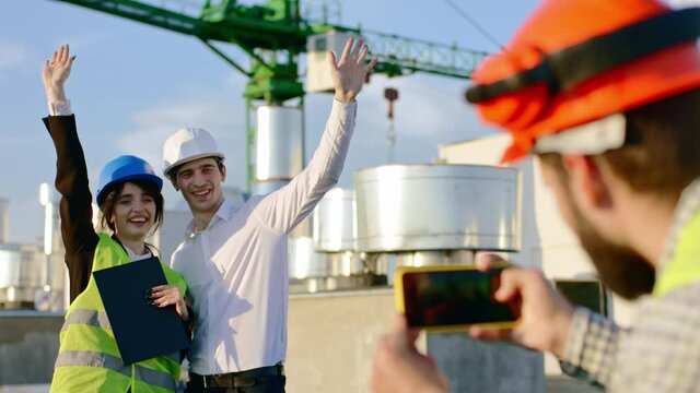 On the top of construction site charismatic guy take pictures for two young engineers lady and man day posing in front of the smartphone wearing safety helmets