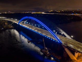 Naklejka premium Aerial drone view. Modern arch cable-stayed bridge in Kiev in the evening. Colored lighting of the arch of the bridge.
