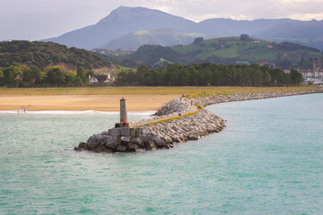 Coast with lighthouse and moor with town and mountains on background. Biscayne bay with scenic coastline. Basque country landscape. Breakwater stones and lighthouse. Commercial dock.