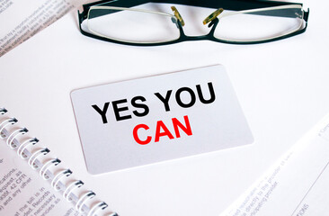 Text Yes You Can on a business card lying on a notepad with eyeglasses and text documents