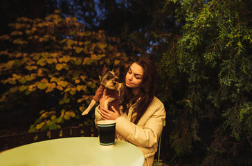 Beautiful woman sitting in a cafe on the street in the evening with a cup of coffee and a dog in her arms. Leisure evening with pet terrace.