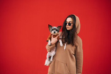 Portrait of a stylish lady with a dog in her arms on a background of a red wall, looking away at a pet. Isolated.