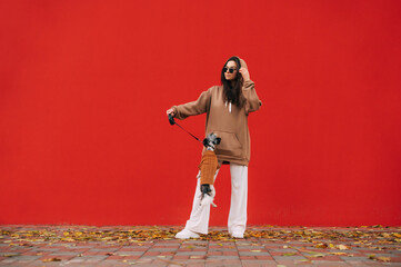 Stylish female owner plays with a small dog on a background of a red wall, the pet jumps, the lady looks away and adjusts her sunglasses.
