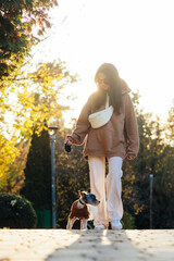 Fototapeta premium Attractive lady stands in a beautiful park at sunset with a small dog in her arms and hugs her pet and smiles.
