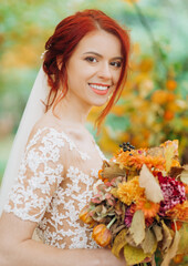 Amazing red-haired bride stands in autumn park with a bouquet of flowers in her hands.