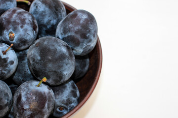 Several ripe blue plums in a deep clay plate on a white background with a copy space