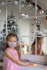 New year in coronavirus conditions. A little girl in a mask on her face in a new year's interior. Light girl in pink dress and the mirror. Beautiful Christmas decorations