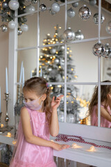 New year in coronavirus conditions. A little girl in a mask on her face in a new year's interior. Light girl in pink dress and the mirror.