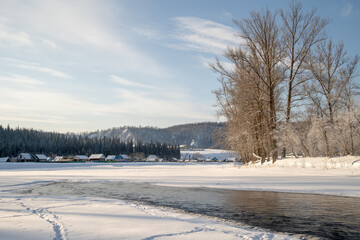 Mountain river on a frosty winter day. On the shore there are trees covered with frost and a village.