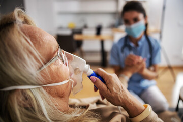 Closeup of ill blond senior woman sitting at home and taking oxygen from respirator while nurse with protective mask on sitting next to her and holding her hand during corona virus. - obrazy, fototapety, plakaty