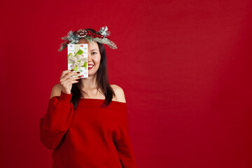 mock-up of a happy Asian young woman in a Christmas wreath hiding half her face behind a gift box, on a red background.