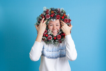 a laughing Asian young woman looks through a Christmas wreath of Christmas tree twigs, cones and ornaments, isolated on a blue background.