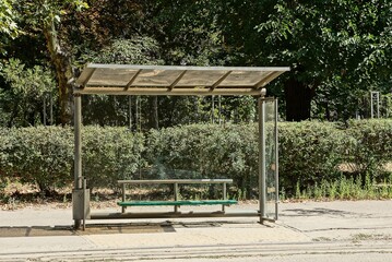 one empty transparent glass bus stop with a bench on the asphalt by the road against a background of green vegetation