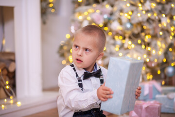 Christmas tree, a boy with Down syndrome sits on the floor and holds a box with a gift in his...