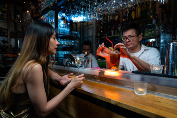 Professional male mixologist bartender preparing mixed alcoholic cocktail drink serving to woman sitting at bar counter in nightclub. Asian barman making mixed liquor into decorative cocktail glass.