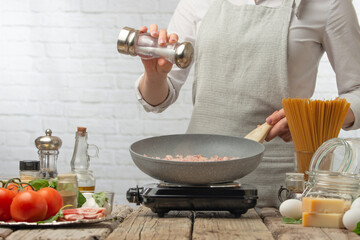 Professional chef in white uniform salts bacon in pan for cooking pasta alla carbonara. Backstage of preparing traditional italian dish on white background. Recipe book.Cooking process concept.