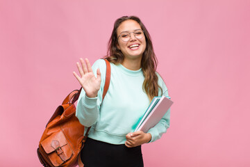 young hispanic woman smiling happily and cheerfully, waving hand, welcoming and greeting you, or...