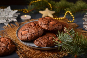 Chocolate cookies with almonds on a gray plate, on a sacking, in the distance a fir branch and a garland.