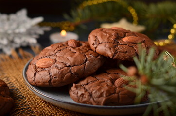 Chocolate cookies with almonds on a gray plate, on a sacking, in the distance a fir branch and a garland.