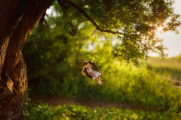Happy little girl swinging on a swing on the branches of a large tree in the rays of the setting sun.Happy childhood concept