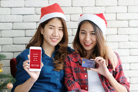 Two young cute asia women holding mobile phone with Christmas discount coupon and credit card for shopping online, e commerce, holiday shopping online