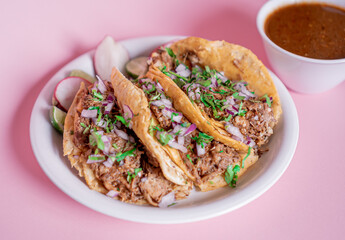 Delicious tacos of birria on a white plate with a broth in the back.