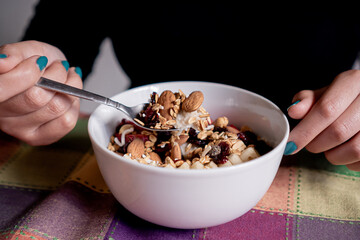 Person eating homemade oatmeal with banana, strawberries, granola, blueberry, coconut and almonds with a spoon inside a white plate.