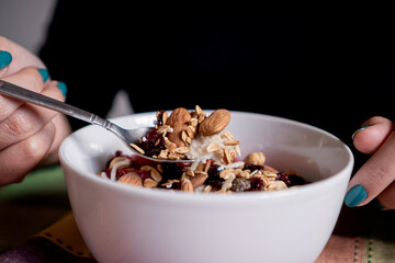 Person eating homemade oatmeal with banana, strawberries, granola, blueberry, coconut and almonds with a spoon inside a white plate.
