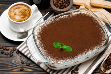 Traditional Italian Tiramisu dessert in glass baking dish, coffee grinder, savoiardi cookies and cup of fresh hot espresso coffee on wooden background