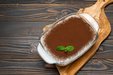 Traditional Italian Tiramisu dessert in glass baking dish on cutting serving board on wooden background