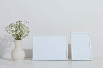 A vase with flowers and a white photo frame on the table. Light stylish background. Preparation for inscriptions, copy space.