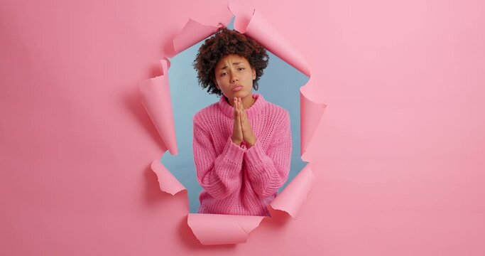 Sad African American woman presses palms to heart makes praying gesture asks for favor has imploring expression looks helpless at camera wears casual clothes poses in ripped hole. Please help