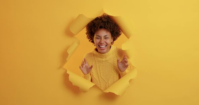 Shocked African American woman suddenly appears in torn paper hole of yellow background raises palms and tries to frighten someone looks gladfully dressed casually. Female tries to be spooky