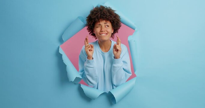 Superstitious hopeful dark skinned woman crosses finger for good luck truly believes in something good happen wishes dream come true poses in paper hole of blue background. God will help me I know