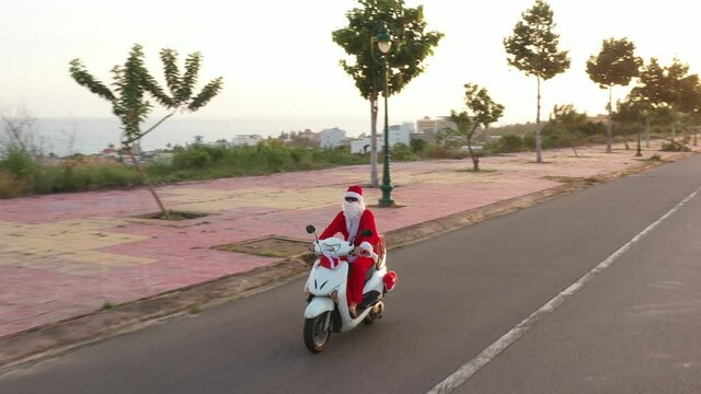 Santa on white motorbike with gift box and gift bag in summer sunset. New year and Christmas content. Boy in Santa costume rides by bike and goes to us. Aerial round view 