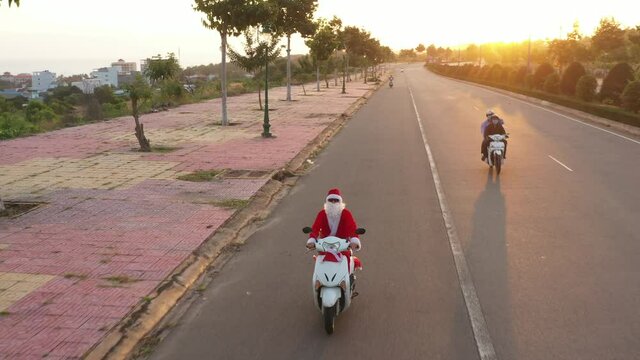 Santa rides a moped along the road with gifts for Christmas and New Year holidays. Cars and motorcycles overtake him. Christmas in summer, warm New Year. Boy in Santa costume rides by white motorbike