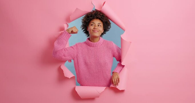 Self confident independent dark skinned woman raises arm and shows her strength looks assured at camera feels powerful wears casual knitted sweater models through ripped hole pink paper background.