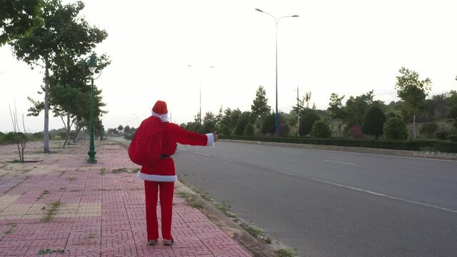 Santa is hitchhiking on the road with gift bag in summer sunset. New year and Christmas content. Boy in a Santa suit stands with his back and catches a ride on the road