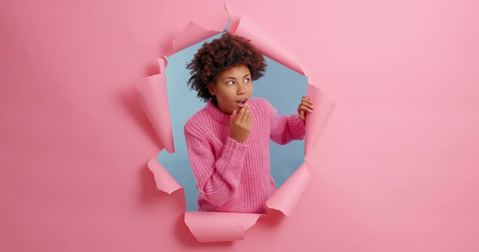 Shocked impressed dark skinned young woman looks away and points at copy space shows breathtaking advertisement smiles happily dressed in knitted jumper poses over torn pink paper background.