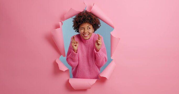 Cheerful smiling Afro American woman waits for special moment keeps fingers crossed makes wish smiles broadly gestures with anticipation wears sweater poses in ripped hole. Wishing good luck